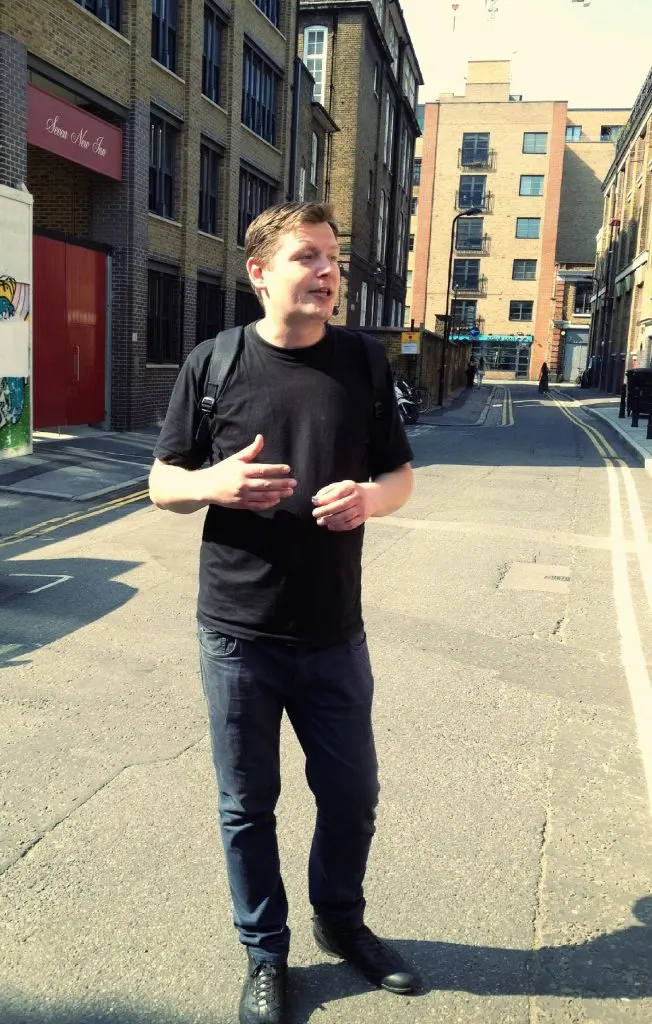 A blonde man wearing a black t-shirt and jeans leading a tour in London 