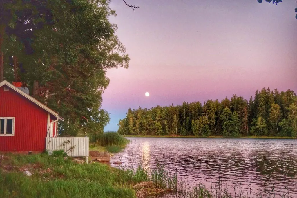 Midnight sun over a river lined by trees and a small red cabin