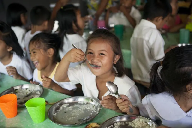 A young girl eating as she smiles to the camera, and other children sitting at the table eating and laughing 