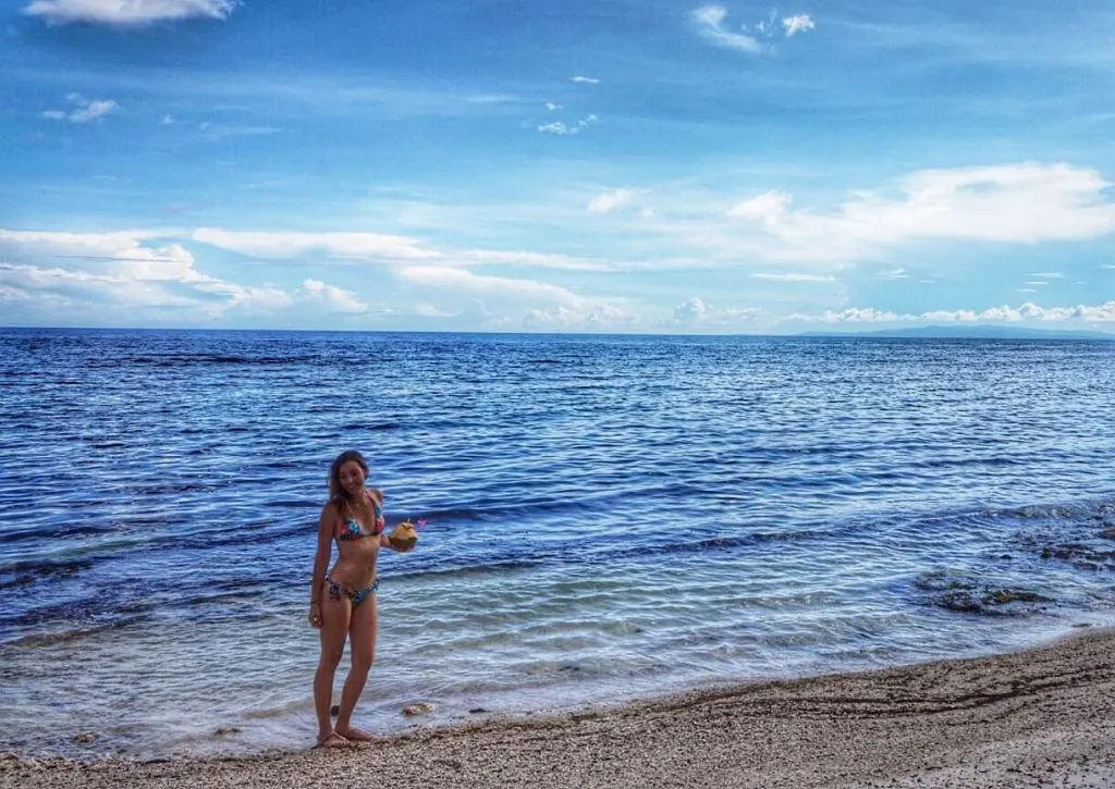 A woman in a bikini standing on the shores of the sea and holding a coconut