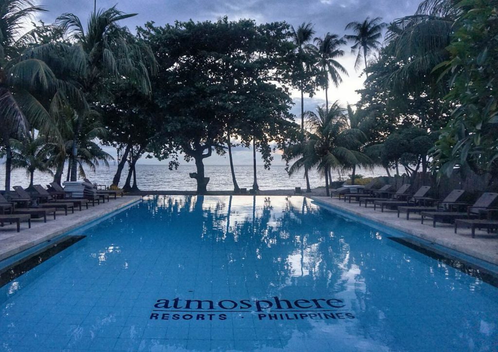 A large swimming pool with the words ''Atmosphere Resorts Philippines'' written in the bottom, surrounded by palm trees and overlooking the sea