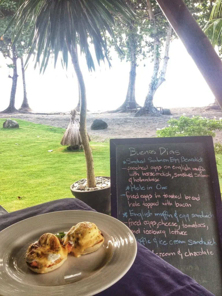 A plate with eggs benedict set in a table outside, next to a board that has the breakfast menu written