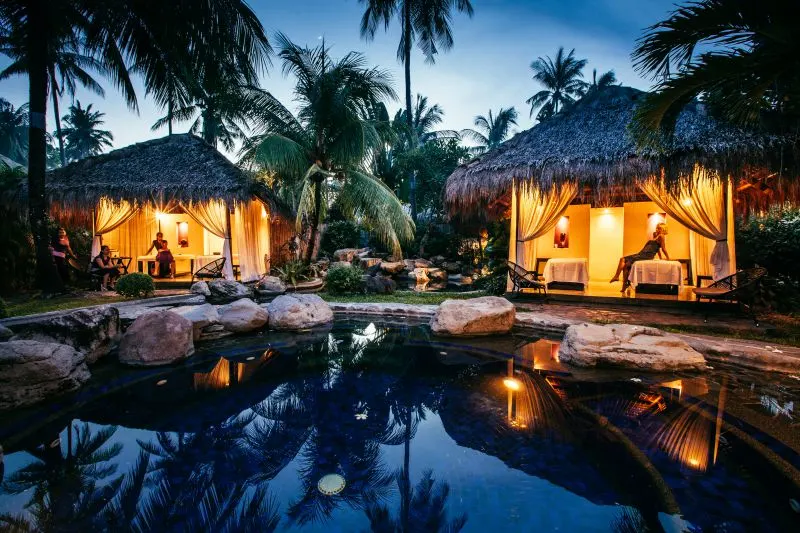 Two spa cabins lit up with warm lights set next to a pool and surrounded by palm trees