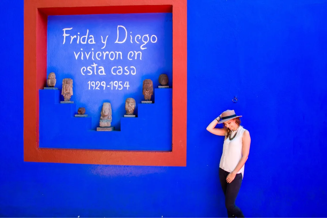 A woman standing in front of a blue wall at Frida Kahlo's house with a whtie sign that reads, in Spanish, Frida and Diego lived in this house 1929 - 1954