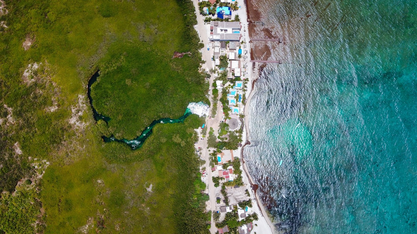 An overhead shot of the Caribbean Sea on the right, a long stretch of buildings, and green vegetation on the left