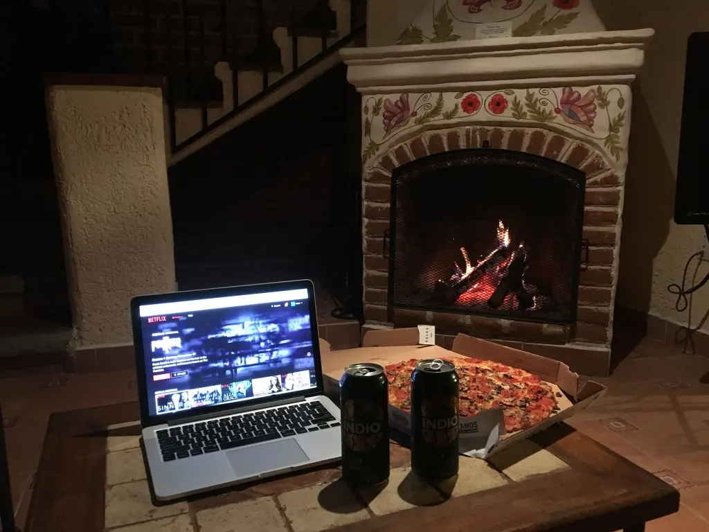 A laptop with a movie on, a box of pizza, and two beer cans over a coffee table, and a fireplace in the background