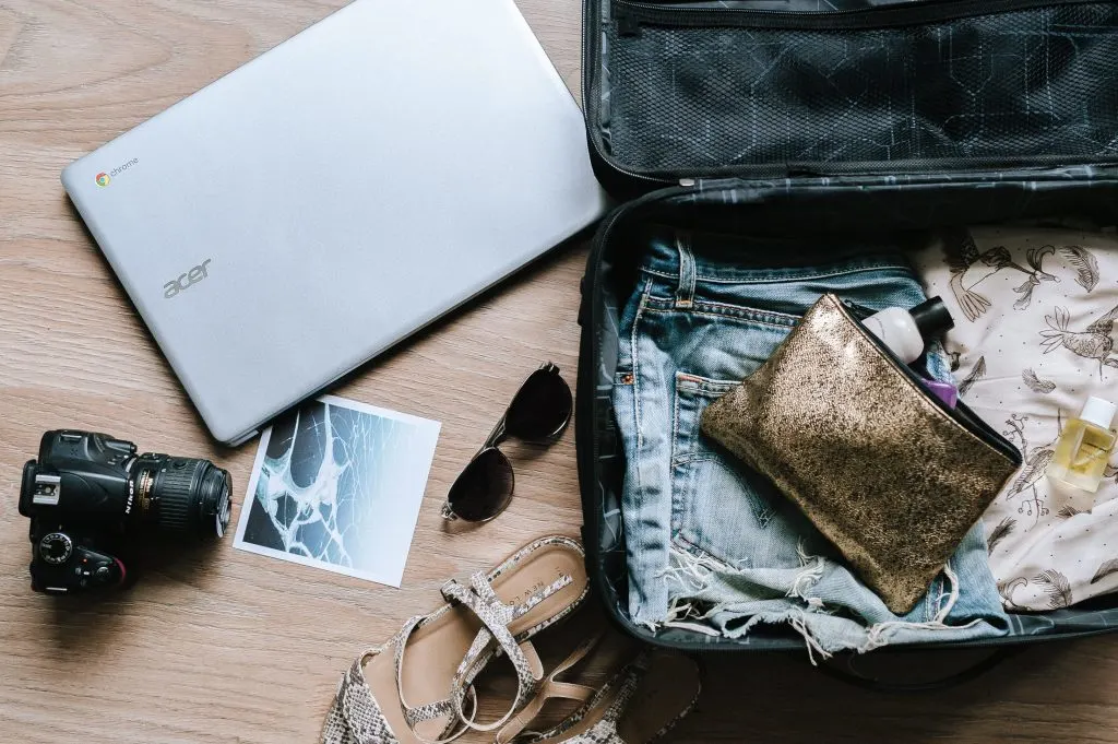 an overhead shot of a carry-on suitcase with a toiletry bag, next to it a pair of sandals, sunglasses, a camera, and a laptop