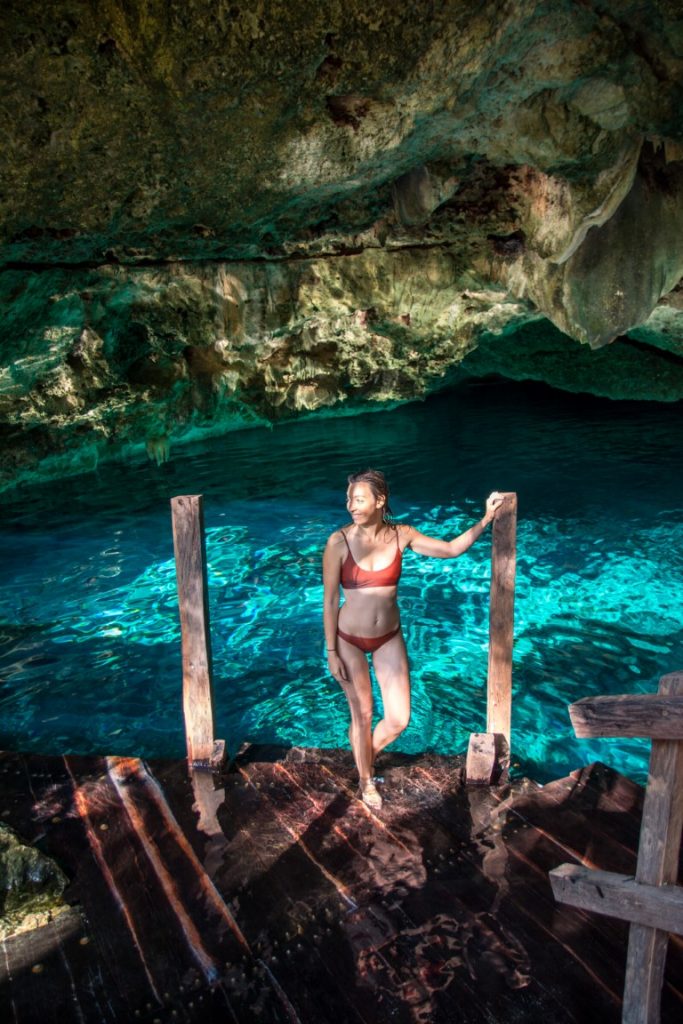 Woman in a brown bikini standing in a cave in front of the electric blue water at Cenote Dos Ojos.