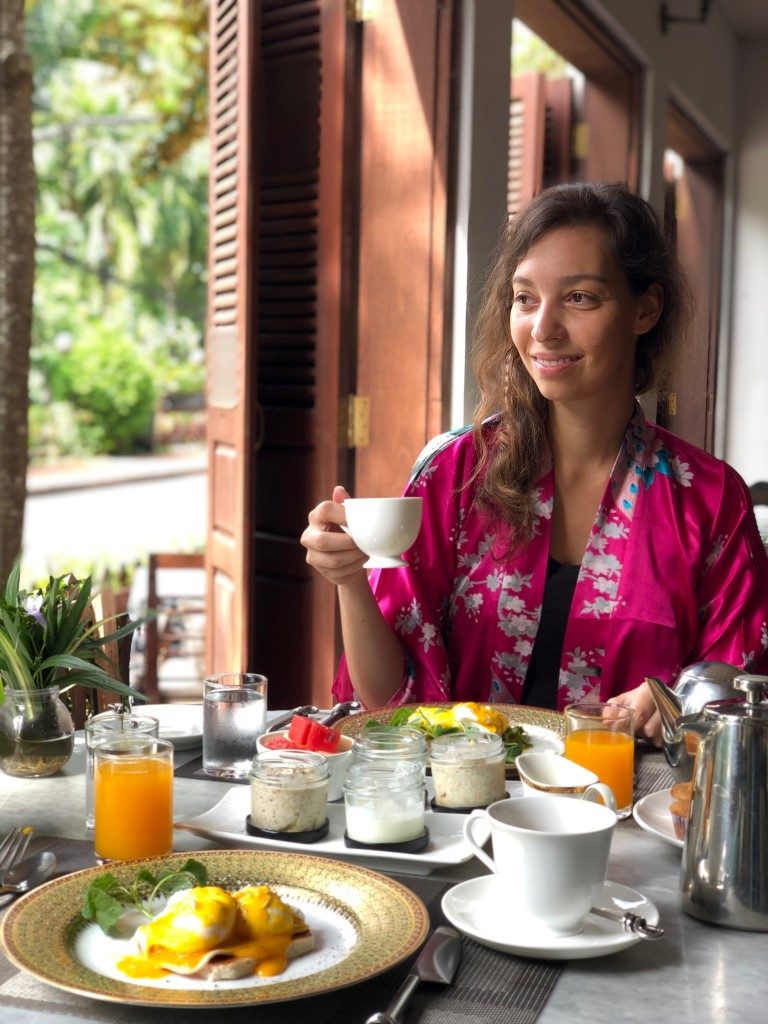 A woman smiling as she holds a coffee mug, sitting in front of a table full of breakfast dishes