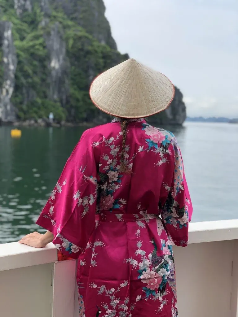 A woman wearing a Vietnamese hat and a silk pink robe admiring the views from a boat's balcony