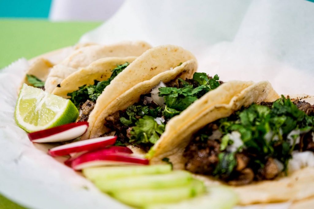 Three Mexican tacos served on a plate, with lemon wedges on the side