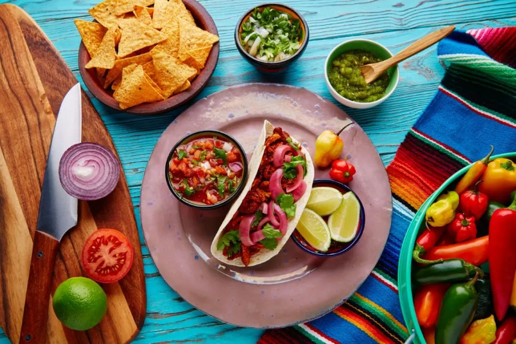 A kitchen table with a pink plate in the center with a Mexican taco, and bowls with lemon wedges, guacamole, and chips