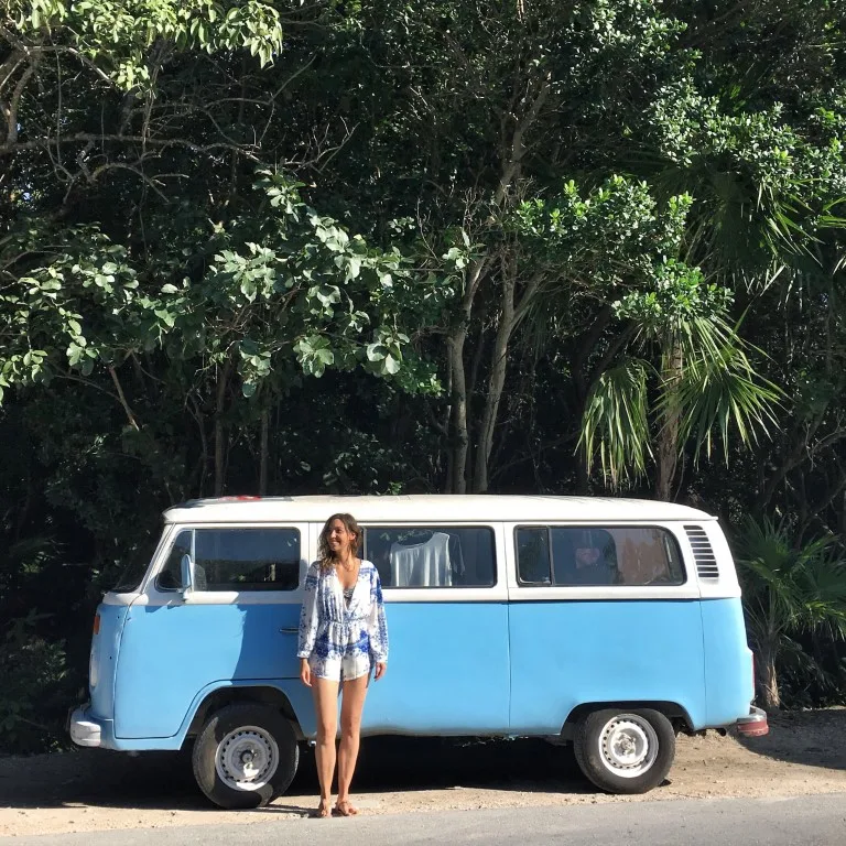 A woman standing in front of a white and blue van, with lush vegetation in the background