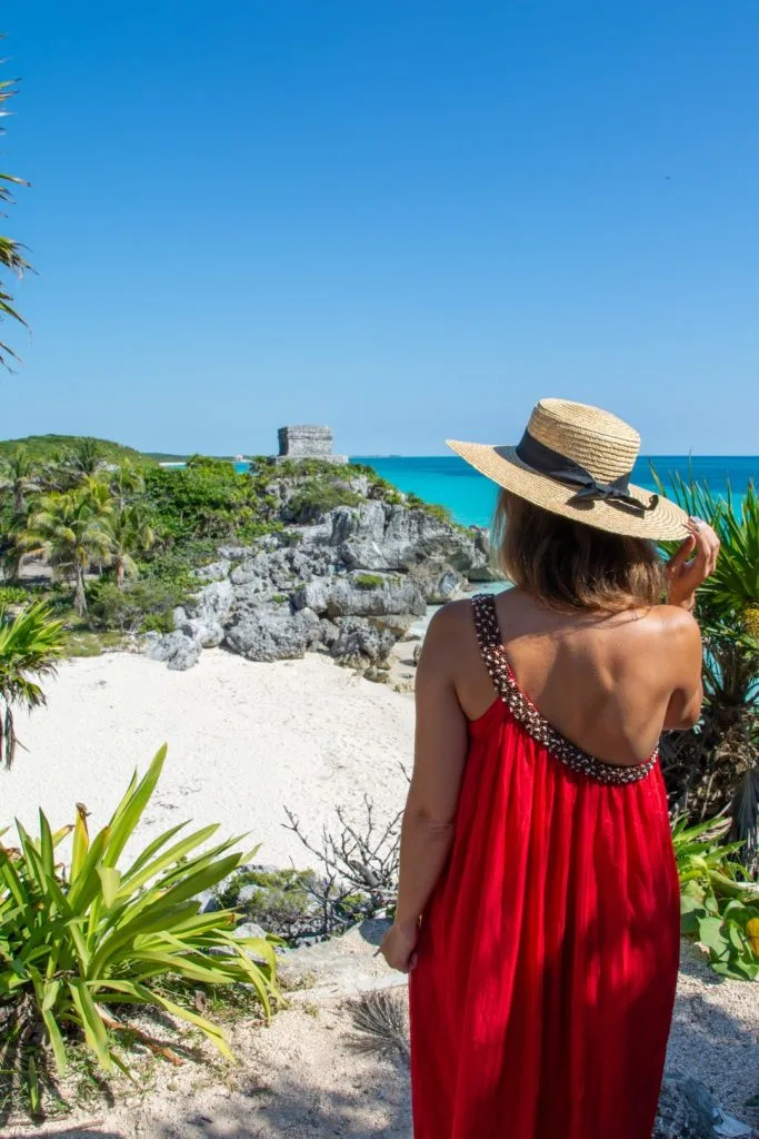 Woman wearing a red dress and a straw hat standing in front of the Tulum ruins.