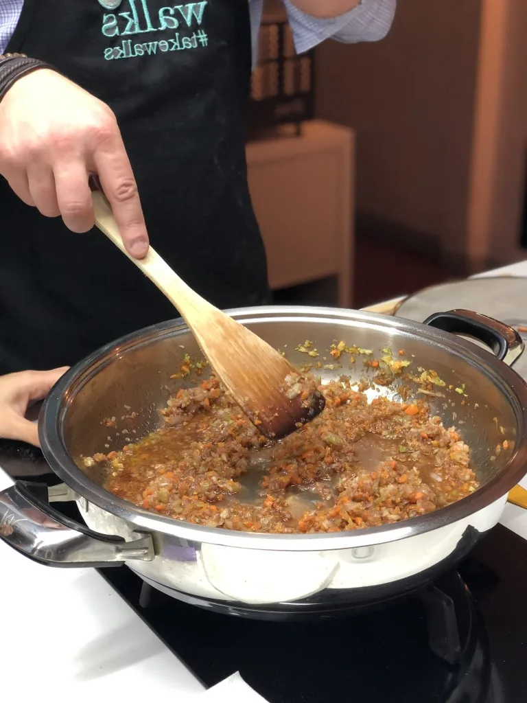 A hand holding a wooden spoon over a pan, sauteing onions and other vegetables 