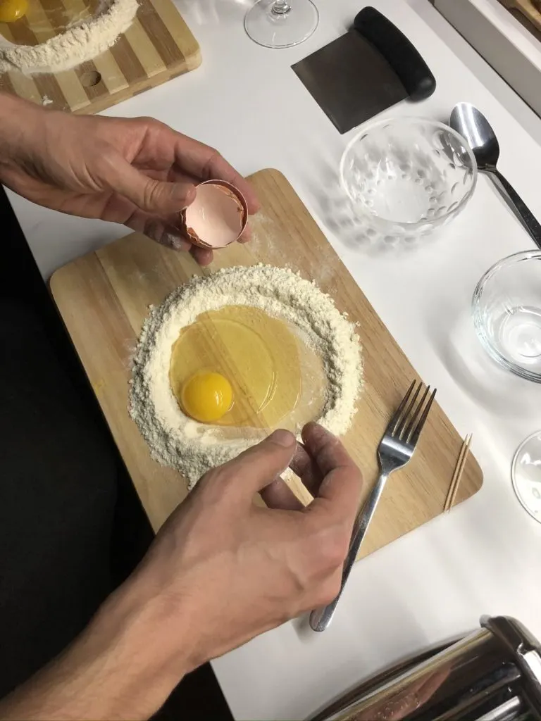 Two hands holding an eggshell over a wooden board, where there's a ring of flour with an egg in the center 