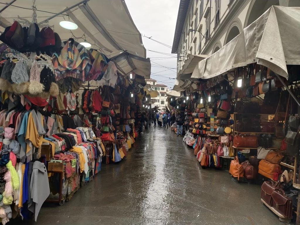 Mercato di San Lorenzo, with colorful clothes and handbags on both sides of a passageway that's wet from the rain 