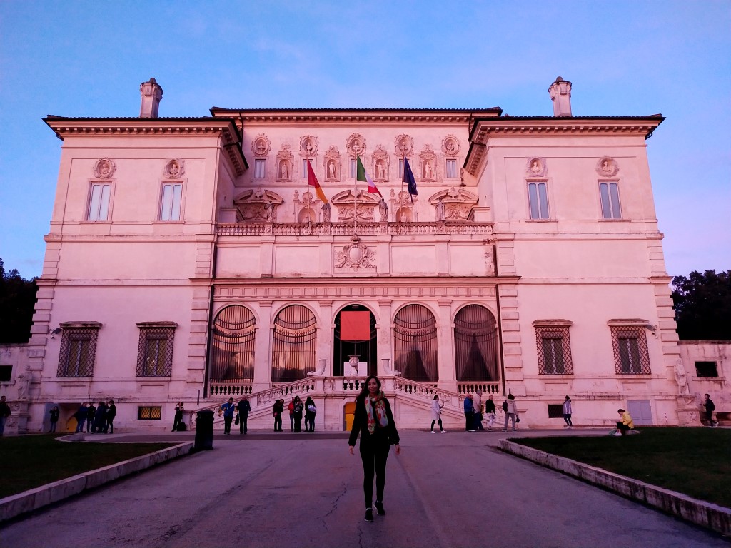 A girl standing in front of Galleria Borghese, which is tainted pink because of the light 