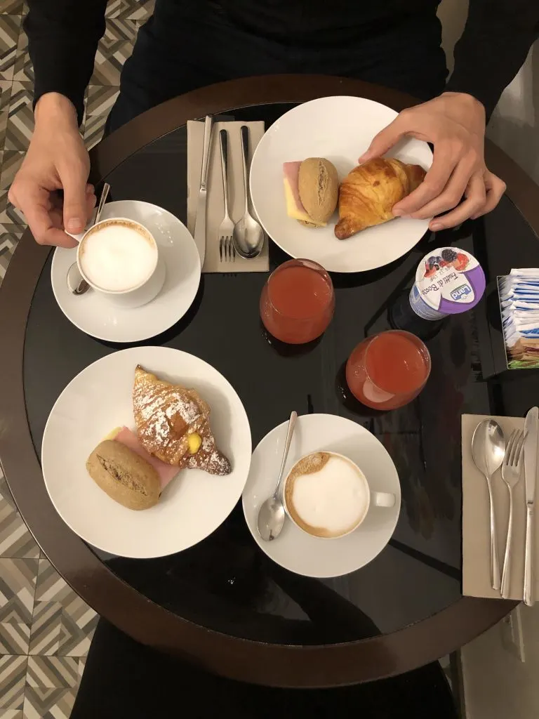 A breakfast table seen from above, with two coffee mugs, two glasses of juice, and two plates with croissants and a panini