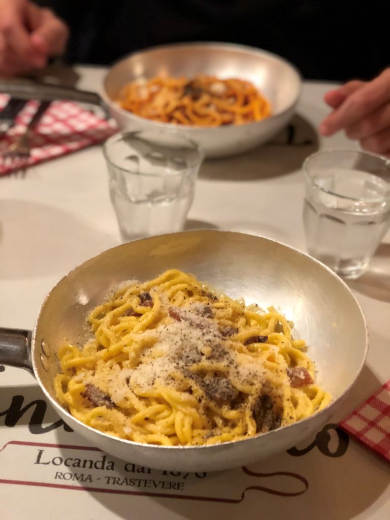 Handmade pasta, seasoned with pepper and cheese, presented in a pan. In the background there's another pan of pasta, and two glasses of water