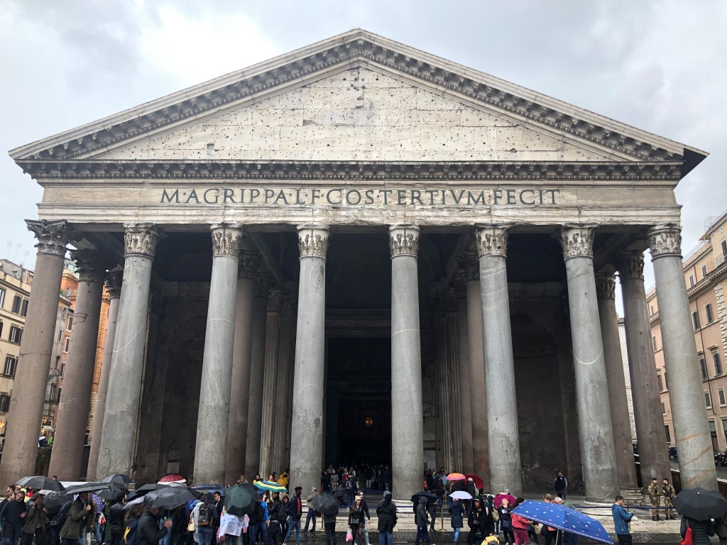 The Pantheon in Rome on a rainy day, with a small crowd holding umbrellas standing in front of it 