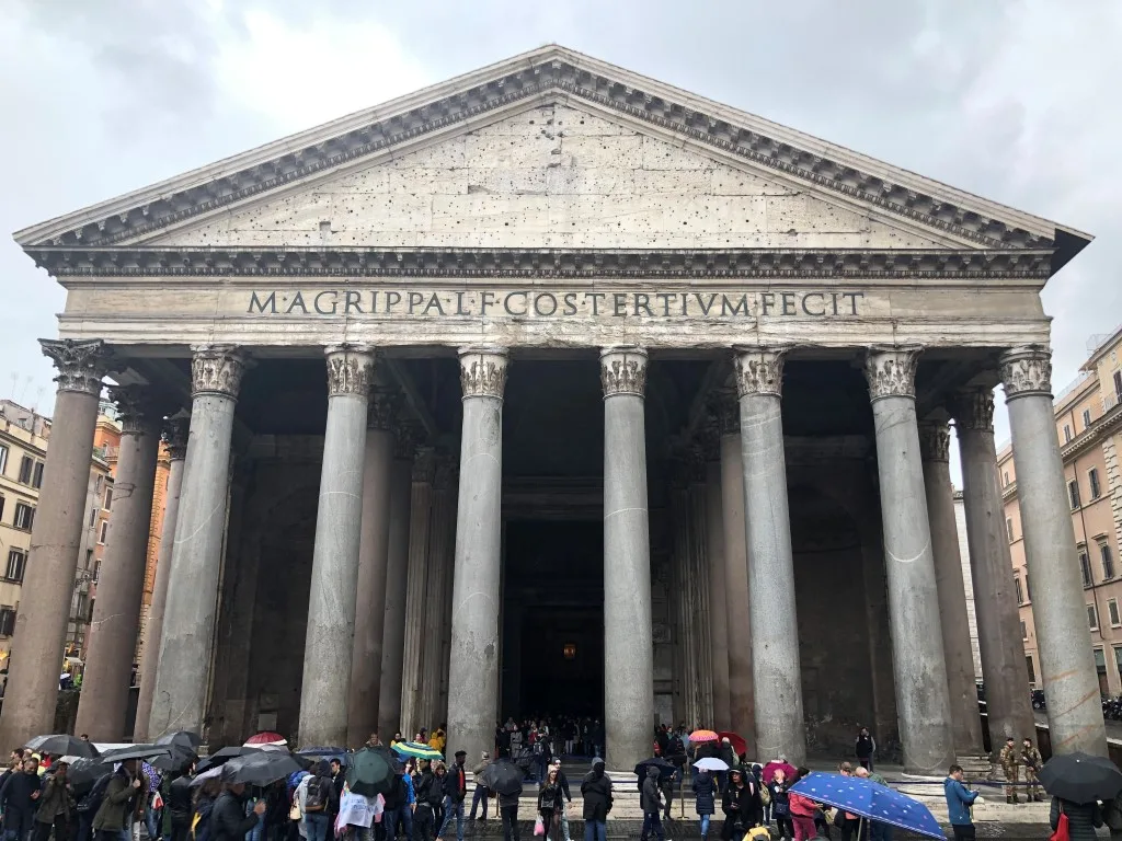 The Pantheon in Rome on a rainy day, with a small crowd holding umbrellas standing in front of it 