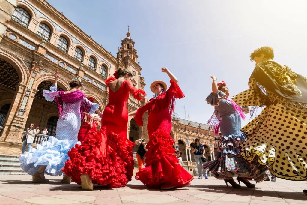 Image of 5 women dressed in red, blue, and yellow flamenco attires dancing in Plaza de España. Watching a flamenco show is one of the best thigns to do in Seville