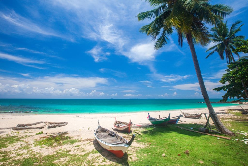 A beach in Thailand with turquoise waters, white sand, and boats under palm trees