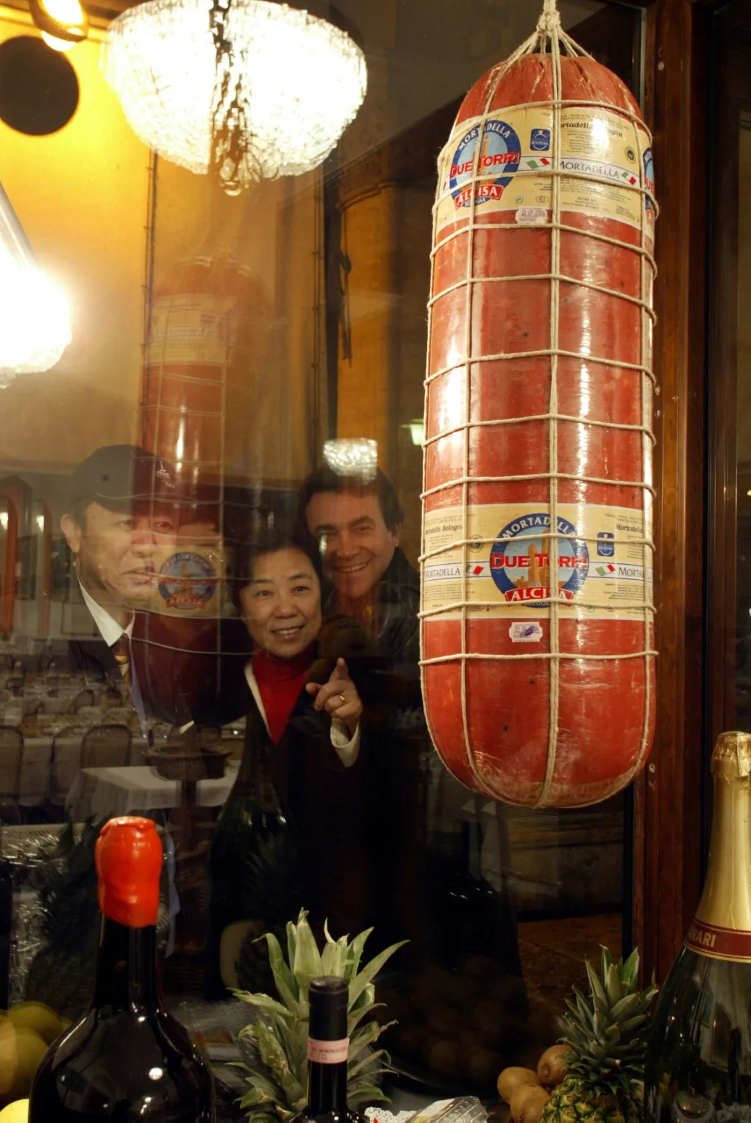 A large mortadella hanging on display, and a group of people admiring it from the window