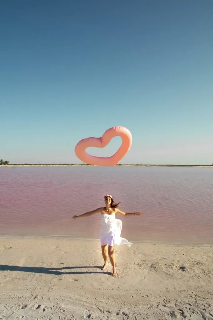 A woman with a white dress playing with a pink inflatbale heart in front of a pink lagoon.