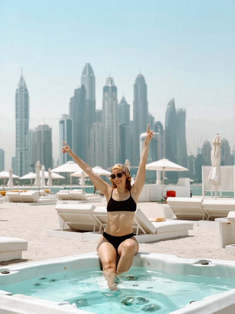 A woman sitting on the edge of a jacuzzi with skyscrapers and sunbeds in the background