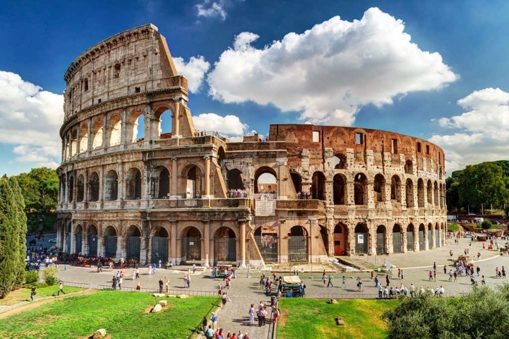 Image of the Colosseum under a blue sky with some clouds, and people walking around it