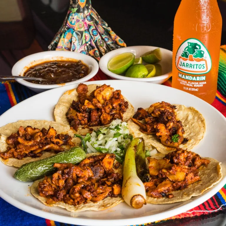 A white plate with five tacos, and in the background a bowl with salsa, another one with lemon wedges, and a small bottle of an orange-flavored soft drink