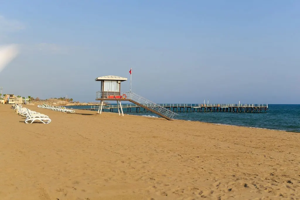 Image of a beach with a pier, white sun chairs, and a lifeguard tower. Chilling in the beach is one of the best things to do in Avila Beach, California, where this picture was taken