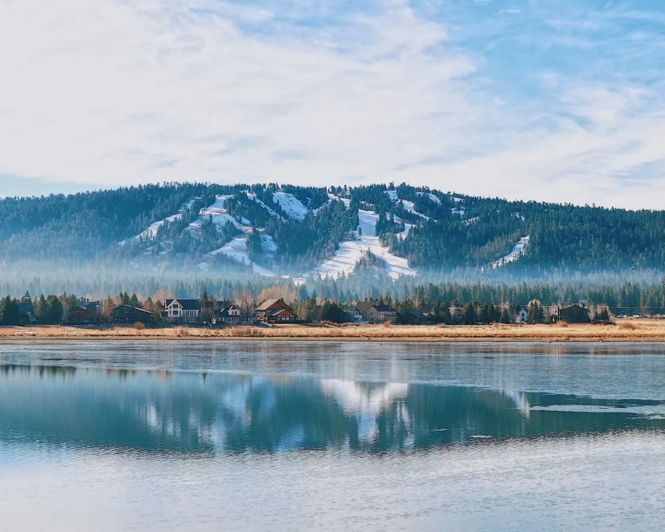 Big Bear Lake, and in the distance a few houses on the lakeside, and a mountain covered in trees and snow 