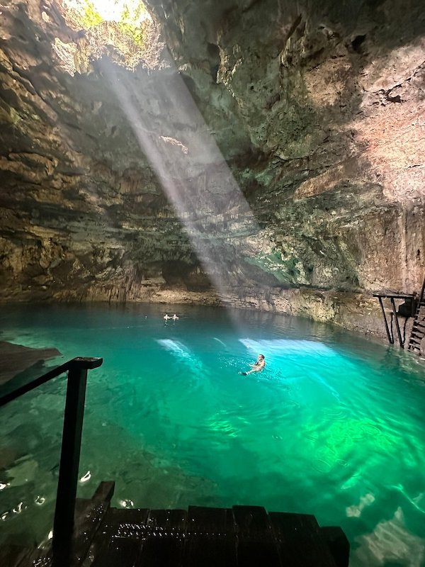 A cave cenote with turquoise water and limestone walls, and a ray of light entering from the top. One person is swimming.