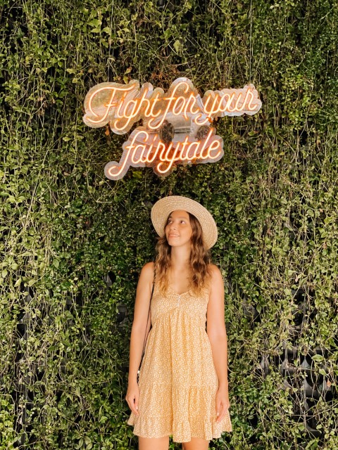 A woman in a yellow dress standing in front of a green wall with a neon sign that reads Fight for your Fairytale