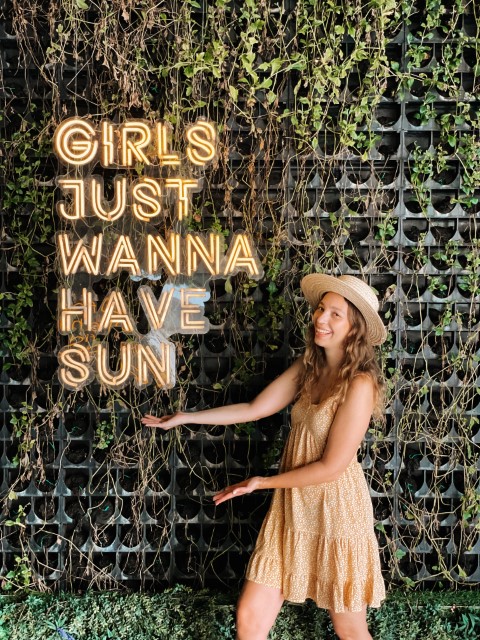 A woman in a yellow dress and a hat smiling next to a neon sign that reads Girls Just Wanna Have Sun