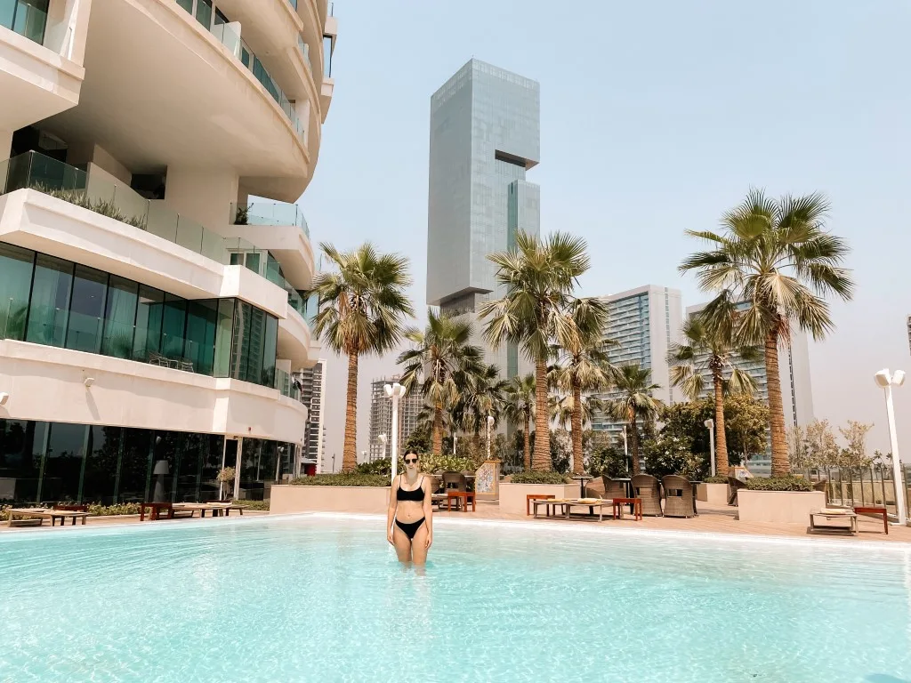 A woman in a black bikini standing in a swimming pool, with buildings and palm trees in the background