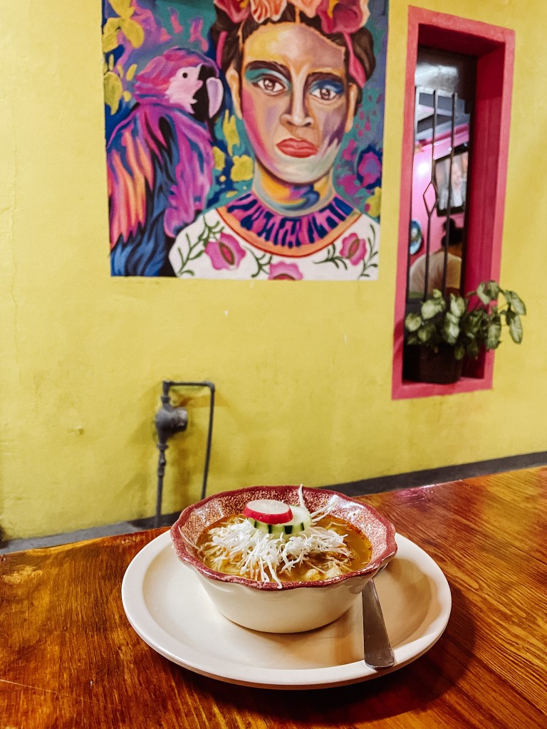 A dish of traditional Mexican cuisine set on a table, and a painting of Frida Kahlo in the background