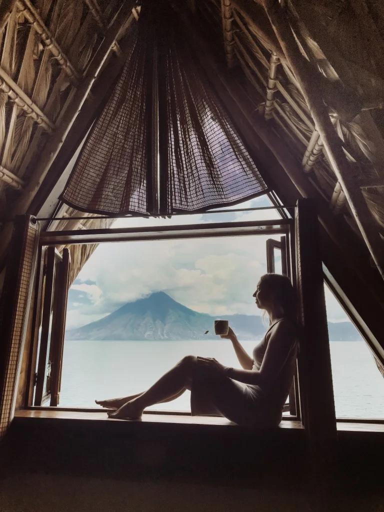 A woman having coffee on the windowsill of a rood, overlooking the lake and volcano outside