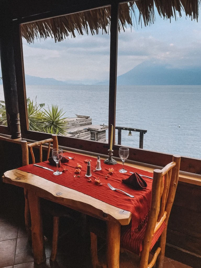 A dinner table set for two people next to a large window that looks out to a lake and volcanoes 