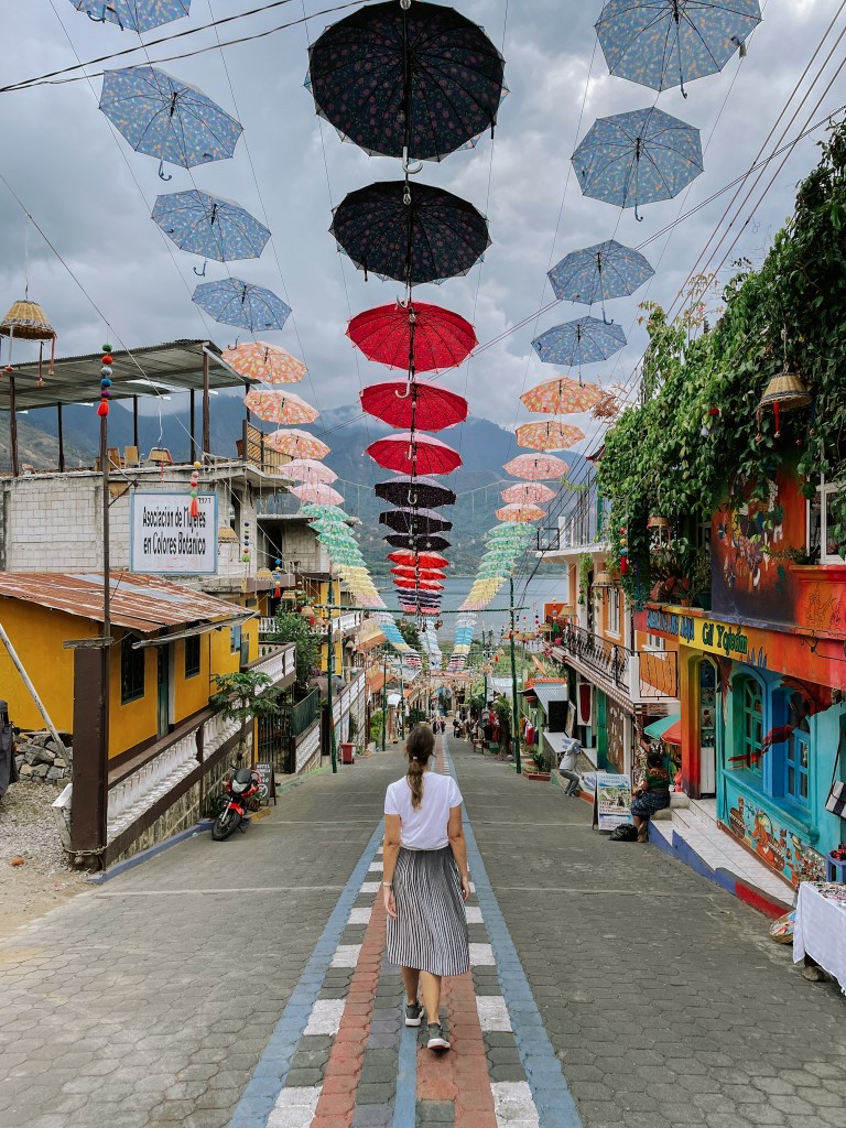 A woman walking down a cobblestone street lined by colorful buildings, and umbrellas hanging above her