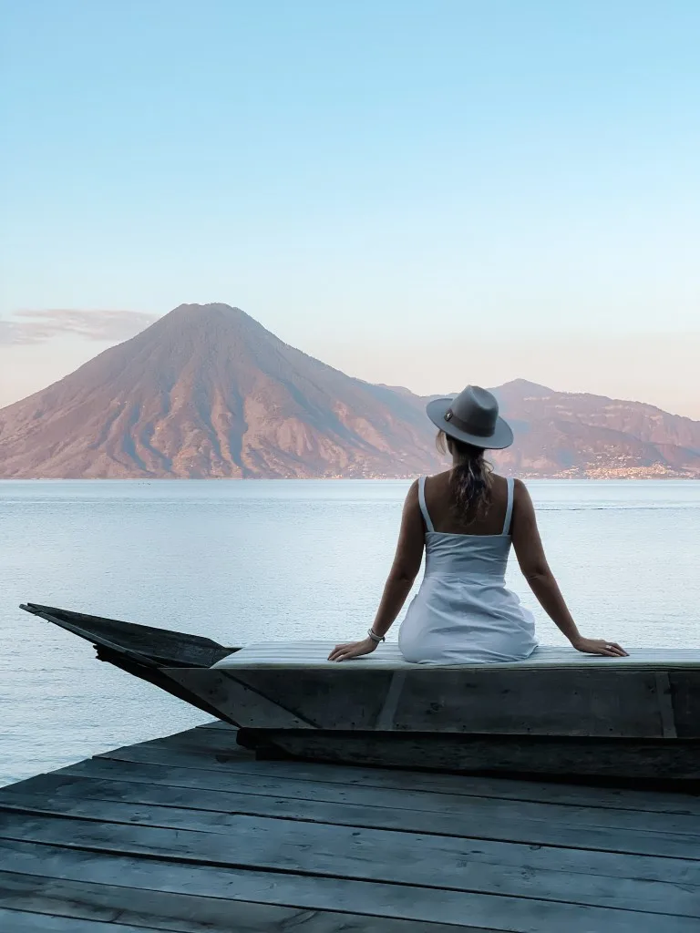 A woman sitting on the shores of a lake, with a volcano in the background