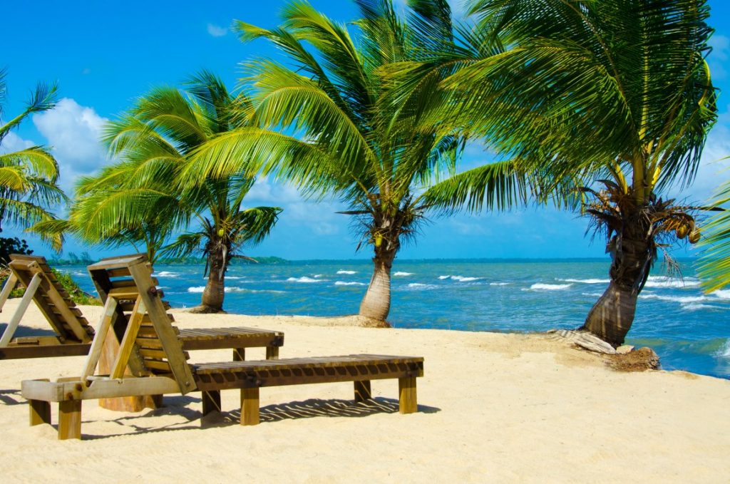 A tropical beach with palm trees swaying in the wind, wooden beach loungers, and the blue sea inserted in a post about going from Guatemala to Belize 