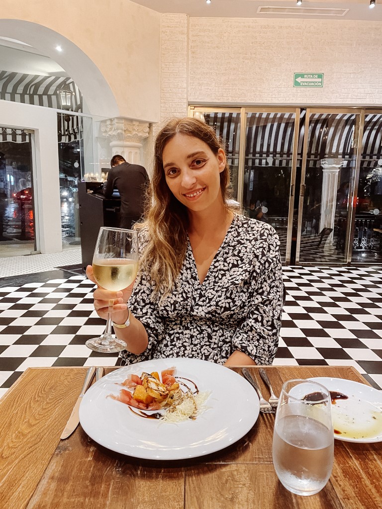 A woman sitting in a restaurant, with a dish in front of her as she holds a glass of white wine and smiles for the camera