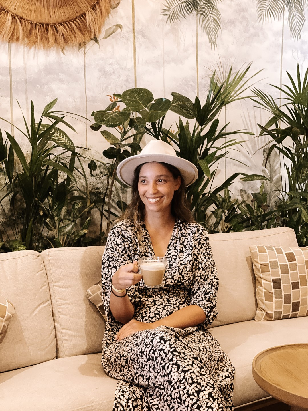 A woman sitting on a couch, smiling as she drinks a latte