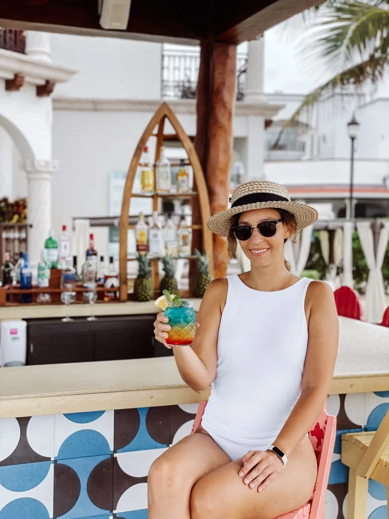 A woman in a white swimsuit, sitting at a beach bar with a cocktail in hand