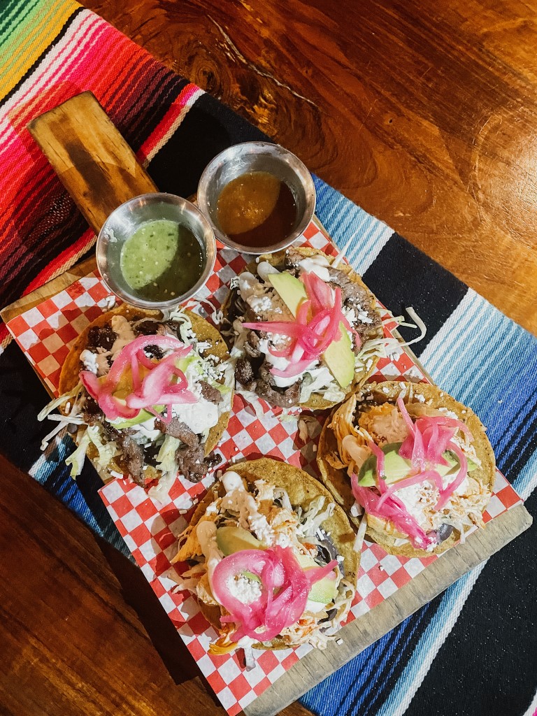 A wooden board with a set of four tacos and two small bowls of salsa