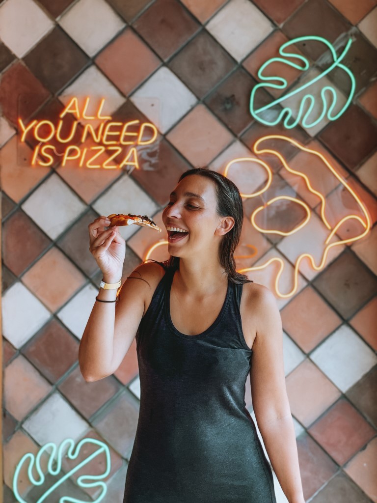 A woman eating a slice of pizza in front of a wall with neon signs that read ''all you need is pizza''